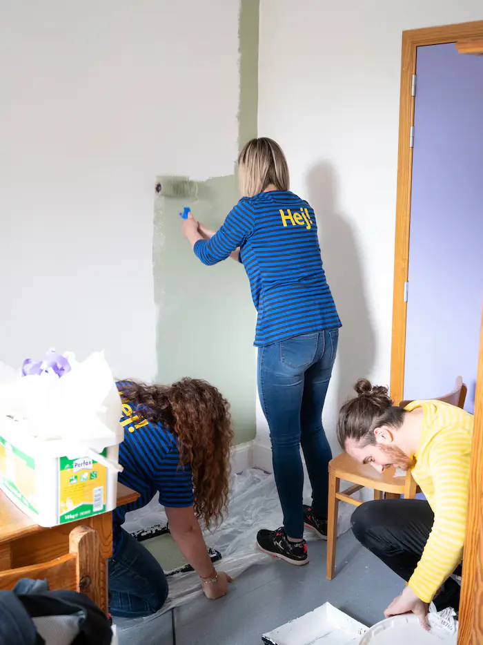 IKEA employees during volunteering activity day. The woman wearing a blue shirt is painting wall in the colour green. While two workers are squatting on the floor, and help to prepare the room for painting.